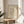May include: Framed beige poster with black line drawing of woman's face and dove, titled "PICASSO" at top and "MUSÉE D'ORSAY" at bottom, displayed on round wooden stool next to beige upholstered chair with white pillow and green plant leaves visible, natural sunlight creating soft shadows on neutral wall background