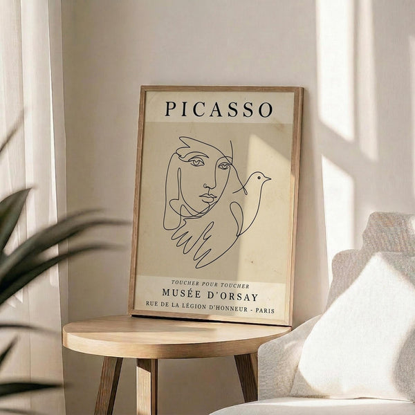 May include: Framed beige poster with black line drawing of woman's face and dove, titled "PICASSO" at top and "MUSÉE D'ORSAY" at bottom, displayed on round wooden stool next to beige upholstered chair with white pillow and green plant leaves visible, natural sunlight creating soft shadows on neutral wall background
