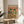 May include: Framed poster showing two cartoon frogs wearing sombreros and sunglasses at a Mexican feast with tacos, margaritas, and colorful piñatas. The artwork features vibrant orange background with multicolored "FROG FIESTA" text. Natural wood frame sits on round wooden side table next to beige upholstered chair with white throw pillow. Soft natural lighting creates shadows on white wall, with plant leaves visible in corner.