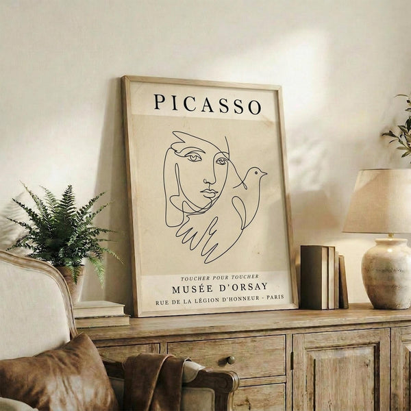 May include: Framed Picasso-style line drawing poster displayed on rustic wooden console table alongside fern plant, books, and ceramic vase. The minimalist artwork features continuous line portrait of woman with dove in beige tones with black text referencing Musée d'Orsay Paris. Natural lighting creates warm shadows on cream walls in cozy living room setting with brown leather furniture visible.