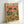 May include: Framed poster print leaning against white wall showing two cartoon frogs wearing sunglasses and Mexican hats sitting at table with tacos, margaritas, guacamole, chips, and tequila bottles. Orange sunset background with colorful piñatas. One frog wears striped poncho, other has bow tie. Wooden frame, vibrant colors with festive Mexican party theme.