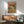 May include: Framed poster displaying two cartoon frogs in Mexican attire sharing a festive meal. One frog wears a sombrero and colorful striped poncho, the other sports a fedora and bow tie. They sit at a table laden with tacos, guacamole, and tequila bottles against an orange background with hanging piñatas. The wooden-framed print hangs above a mid-century modern walnut credenza with a white ceramic lamp, positioned near a mint green upholstered armchair with wooden arms.