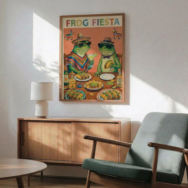 May include: Framed poster displaying two cartoon frogs in Mexican attire sharing a festive meal. One frog wears a sombrero and colorful striped poncho, the other sports a fedora and bow tie. They sit at a table laden with tacos, guacamole, and tequila bottles against an orange background with hanging piñatas. The wooden-framed print hangs above a mid-century modern walnut credenza with a white ceramic lamp, positioned near a mint green upholstered armchair with wooden arms.