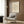 May include: Beige and cream vintage-style Picasso poster showing minimalist line drawing of woman's face with bird and hands, displayed in light wood frame on warm brown wood paneled wall above modern cream armchair with geometric patterned pillow, brass floor lamp and dark round side table with open book nearby