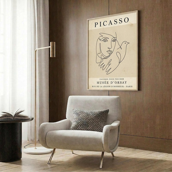 May include: Beige and cream vintage-style Picasso poster showing minimalist line drawing of woman's face with bird and hands, displayed in light wood frame on warm brown wood paneled wall above modern cream armchair with geometric patterned pillow, brass floor lamp and dark round side table with open book nearby