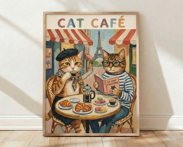 May include: Framed vintage-style poster showing two anthropomorphic cats at a Parisian café table. One orange tabby wears a black beret and scarf while sipping coffee, the other tabby in striped shirt and glasses reads a book titled "Le Chat Philosophe." Table features croissants, macarons, and French press with Eiffel Tower visible in background through café windows with red striped awnings. Artwork displayed in light wood frame against white wall on wooden floor.