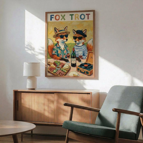 May include: Framed poster showing two cartoon foxes in sunglasses enjoying wine and snacks, with "FOX TROT" in colorful letters at top. One fox wears a tropical shirt and hat, the other a striped shirt and beret. Scene includes charcuterie board, wine bottle, and vintage record player. Poster hangs above mid-century modern wooden sideboard with white lamp, next to teal upholstered armchair in bright, naturally lit room.