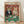 May include: Framed vintage-style poster showing two anthropomorphic cats in formal attire at a green casino table. Tabby cat in black tuxedo with cigar sits beside orange cat in sparkly dress holding playing cards. Table features poker chips, wine bottle, glass, and scattered cards against ornate red wallpaper background. Colorful "CAT CASINO" text spans the top. Light wood frame leans against white wall on wooden floor.