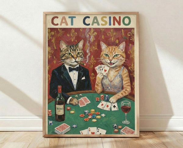 May include: Framed vintage-style poster showing two anthropomorphic cats in formal attire at a green casino table. Tabby cat in black tuxedo with cigar sits beside orange cat in sparkly dress holding playing cards. Table features poker chips, wine bottle, glass, and scattered cards against ornate red wallpaper background. Colorful "CAT CASINO" text spans the top. Light wood frame leans against white wall on wooden floor.