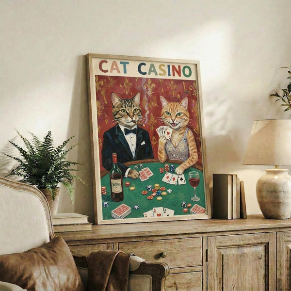 May include: Framed vintage-style poster showing two cats in formal attire playing poker at green felt table with cards, chips, and wine. Tabby cat in tuxedo and orange cat in sparkly dress against ornate red wallpaper background. Display shows rustic wooden console with books, ceramic vase, fern plant, and beige armchair in cozy living room setting.