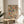 May include: Framed artwork displaying a colorful mosaic of vintage book spines in various sizes, featuring cream, blue, orange, red, and green tones. The print sits on a round wooden side table next to a beige upholstered chair in a bright, minimalist living room with white walls and natural sunlight streaming through windows. A plant's leaves are visible in the corner.