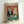May include: Framed vintage-style poster showing two anthropomorphic cats in formal attire at a green poker table. The tabby cat wears a black tuxedo with bow tie, while the orange cat wears a sparkly dress and holds playing cards. Red ornate wallpaper background, casino chips, wine bottle and glass, scattered playing cards on felt table. Light wooden frame leaning against white wall.