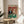May include: Framed poster showing two cats dressed as gamblers at a casino table - tabby cat in black tuxedo and orange cat in sparkly dress, holding playing cards with poker chips and wine bottle on green felt table. Vintage-style artwork with red fleur-de-lis wallpaper background, displayed on round wooden side table next to beige upholstered chair in bright living room with white walls and plant shadows.