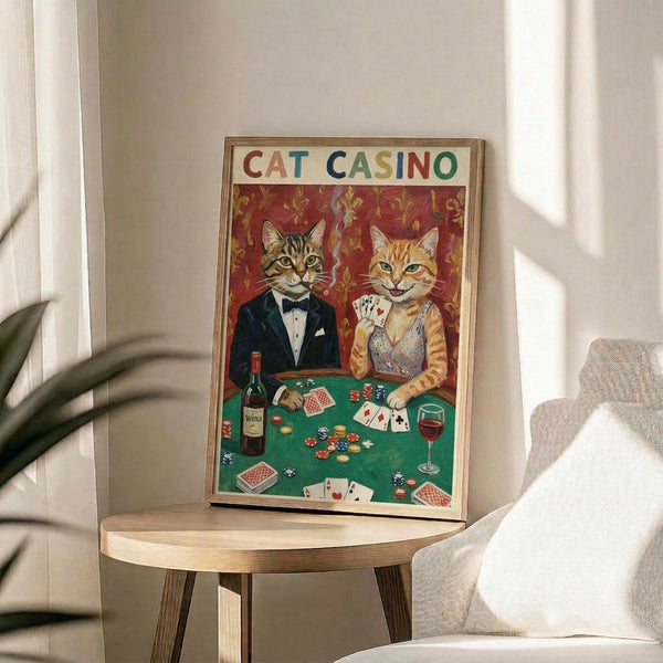 May include: Framed poster showing two cats dressed as gamblers at a casino table - tabby cat in black tuxedo and orange cat in sparkly dress, holding playing cards with poker chips and wine bottle on green felt table. Vintage-style artwork with red fleur-de-lis wallpaper background, displayed on round wooden side table next to beige upholstered chair in bright living room with white walls and plant shadows.