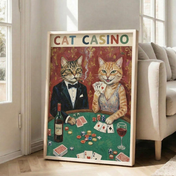 May include: Framed poster print leaning against white wall in bright living room with beige sofa. Artwork shows two cats in formal attire at green casino table - tabby cat in black tuxedo and orange cat in sparkly dress holding playing cards. Red ornate wallpaper background, poker chips, wine bottle and glass, scattered cards on table. Colorful vintage-style illustration in white frame on hardwood floor.