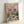 May include: Framed poster print leaning against white wall showing two cats wearing sunglasses having a picnic on checkered blanket. Orange tabby in floral shirt and gray striped cat in blue shirt hold wine glasses. Pink sky with clouds above green grass. Picnic spread includes cheese, grapes, bread, fish, and wine bottle. Colorful illustrated art style in light wood frame.