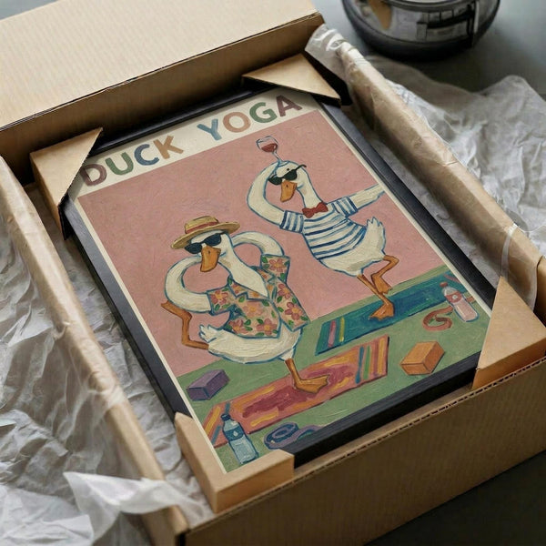 May include: Framed whimsical illustration showing two white ducks doing yoga poses on a pink background, with "DUCK YOGA" text at top. One duck wears a floral shirt and sun hat, the other wears a striped shirt and holds a wine glass. Yoga mat, blocks, and bottle visible below. Print displayed in black frame, being unpacked from cardboard shipping box with protective tissue paper.