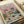 May include: Vintage-style poster print featuring two cool cats in sunglasses enjoying a picnic on a checkered blanket. Orange tabby in floral shirt holds wine glass while gray striped cat sits beside wine bottle. Spread includes cheese, grapes, bread, and fish on plate. Pink sky with fluffy clouds backdrop. Print shown in natural wood frame on white surface with neutral packaging materials visible.
