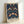 May include: A framed vintage botanical poster displaying an ornate Arts and Crafts pattern with decorative birds, strawberry plants, and flowing foliage in deep navy blue, sage green, coral pink, and cream tones. The design features symmetrical pairs of stylized birds amid intertwining vines, berries, and flowers. Text appears at the bottom identifying the artist and pattern name. The wooden frame sits propped against a white wall.