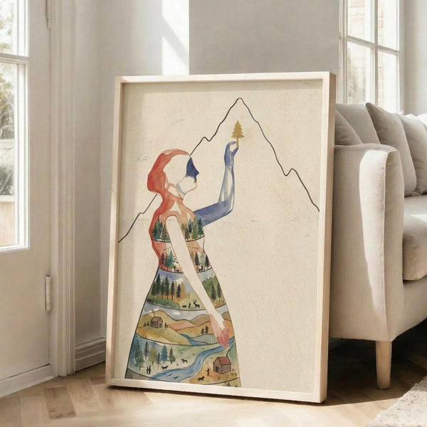 May include: Framed artwork leaning against white wall next to beige sofa in bright room with windows. Print shows watercolor-style woman silhouette filled with landscape scenes including forests, mountains, cabins, and rivers in earthy tones. She holds small tree while reaching toward mountain outline. Light wood frame on hardwood floor.