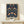 May include: Framed poster featuring William Morris's ornate floral pattern with symmetrical birds, strawberries, and botanical motifs in navy blue, coral, and sage green. The vintage-style print sits on a light wooden shelf beside a small white wrapped gift with natural twine, displayed against a cream wall in natural lighting.