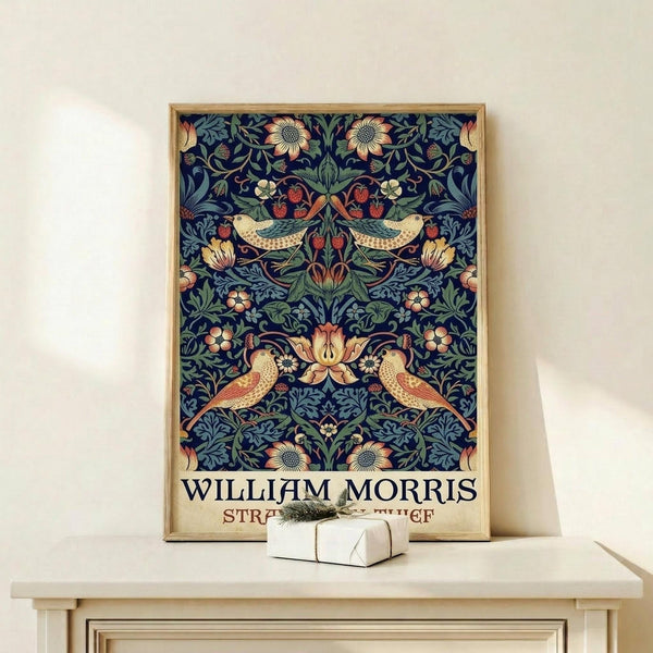 May include: Framed poster featuring William Morris's ornate floral pattern with symmetrical birds, strawberries, and botanical motifs in navy blue, coral, and sage green. The vintage-style print sits on a light wooden shelf beside a small white wrapped gift with natural twine, displayed against a cream wall in natural lighting.