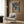 May include: Colorful retro-style poster showing a cat and dog in astronaut suits sharing fried chicken and drinks in space, with planets and stars in background. Poster displayed on wood-paneled wall above modern cream armchair with geometric pillow, next to gold floor lamp and side table with open book.