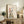 May include: Framed artwork displaying a stylized woman's silhouette filled with a detailed landscape scene featuring mountains, forests, rivers, and small buildings in watercolor style. The piece sits on a rustic wooden console table alongside books, a ceramic vase, and a fern plant, with a beige upholstered chair visible in the foreground in a bright, modern living space.