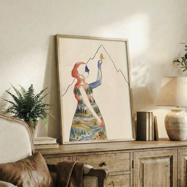 May include: Framed artwork displaying a stylized woman's silhouette filled with a detailed landscape scene featuring mountains, forests, rivers, and small buildings in watercolor style. The piece sits on a rustic wooden console table alongside books, a ceramic vase, and a fern plant, with a beige upholstered chair visible in the foreground in a bright, modern living space.