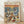 May include: Framed poster print leaning against white wall on wooden floor. Artwork shows two white ducks in sunglasses sitting on yacht deck at sunset, toasting with champagne glasses. One duck wears floral Hawaiian shirt and straw hat, other wears blue striped sailor shirt and white captain's hat. Deck features gourmet food spread including lobster, caviar, cheese, and fruit. Ocean background shows boats and orange sunset sky. Light wood frame surrounds vintage-style illustration.