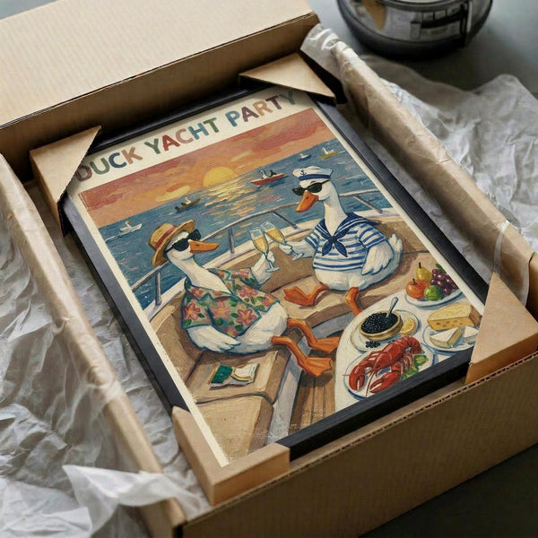 May include: Framed vintage-style poster showing two anthropomorphic ducks on a yacht deck at sunset, one wearing a striped sailor shirt and captain's hat, the other in a floral Hawaiian shirt and sun hat. The scene features a wooden deck with luxury food including caviar, lobster, and wine. The artwork has warm sunset colors with orange sky and blue ocean. The black-framed print is partially unwrapped in a cardboard shipping box with white tissue paper.