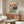 May include: Whimsical illustrated poster showing two dressed cats at a picnic table with gourmet foods, displayed above a mid-century modern walnut credenza in a bright living room with white curved chair, glass coffee table, and warm natural lighting creating soft shadows on neutral walls.