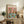 May include: Framed poster print displaying two cartoon cats at a spa table - one wearing cucumber eye mask and towel wrap, another with face mask, surrounded by wine, fruit, spa items, and cheese on turquoise table. Warm wood sideboard setting with books, plant, pottery, and cream chair visible in cozy home interior.