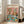 May include: Framed poster print leaning against white wall next to beige sofa in bright living room with large windows. Artwork shows two cats at spa day - one wearing cucumber eye mask holding wine glass, another with face mask and towel turban. Turquoise table displays spa items, fruit plates, cheese, and wine bottle labeled "Purrfection". Colorful retro-style illustration with pink background.