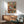 May include: Framed poster showing two anthropomorphic cats on a couch - one orange cat in striped pajamas napping with sleep mask, another cat in suit reading nearby. Colorful "CAT NAP" text at top. Print displayed above modern wooden credenza with white ceramic lamp, next to mid-century sage green upholstered chair in sunlit room with white walls.