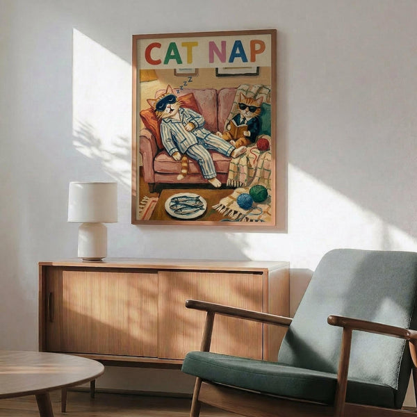 May include: Framed poster showing two anthropomorphic cats on a couch - one orange cat in striped pajamas napping with sleep mask, another cat in suit reading nearby. Colorful "CAT NAP" text at top. Print displayed above modern wooden credenza with white ceramic lamp, next to mid-century sage green upholstered chair in sunlit room with white walls.