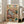 May include: Framed poster print leaning against white wall next to beige sofa in bright living room with large windows. Artwork shows two orange tabby cats in colorful pajamas relaxing on brown armchair - one sleeping with eye mask, one reading book wearing glasses. Whimsical illustration includes yarn balls, fish plate, and cozy home details. Light wood frame, approximately 2-3 feet tall based on furniture scale.