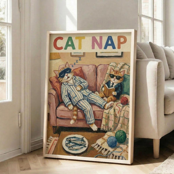 May include: Framed poster print leaning against white wall next to beige sofa in bright living room with large windows. Artwork shows two orange tabby cats in colorful pajamas relaxing on brown armchair - one sleeping with eye mask, one reading book wearing glasses. Whimsical illustration includes yarn balls, fish plate, and cozy home details. Light wood frame, approximately 2-3 feet tall based on furniture scale.