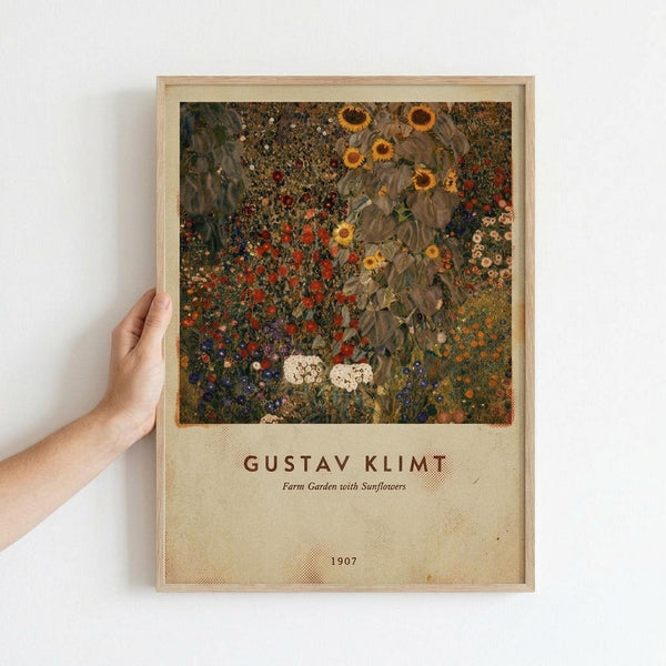 A person holds a framed art print against a white wall. The artwork shows Gustav Klimt's colorful garden painting with vibrant sunflowers, red poppies, and wildflowers in rich yellows, reds, and greens. Below the image is cream-colored text reading "GUSTAV KLIMT" and "Farm Garden with Sunflowers" with "1907" at the bottom. The print has a light wooden frame and vintage-style layout.