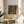 May include: Framed art print of Gustav Klimt's colorful floral garden painting displayed on a round wooden side table next to a beige upholstered chair. The artwork features vibrant sunflowers, wildflowers, and dense foliage in rich greens, reds, and golds with white text below. Natural sunlight streams across the neutral-toned room with white walls and a plant visible in the corner.