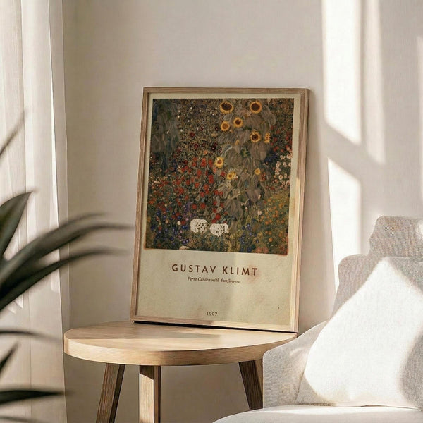 May include: Framed art print of Gustav Klimt's colorful floral garden painting displayed on a round wooden side table next to a beige upholstered chair. The artwork features vibrant sunflowers, wildflowers, and dense foliage in rich greens, reds, and golds with white text below. Natural sunlight streams across the neutral-toned room with white walls and a plant visible in the corner.