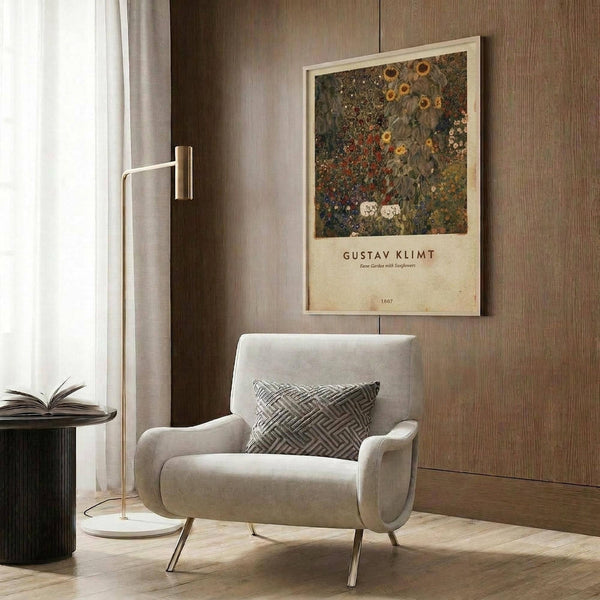 May include: Gustav Klimt art poster featuring a colorful garden scene with sunflowers and floral patterns in rich greens, golds, and reds, displayed in light wooden frame on wood-paneled wall above beige upholstered armchair with geometric patterned pillow, beside modern brass floor lamp and dark round side table with open book