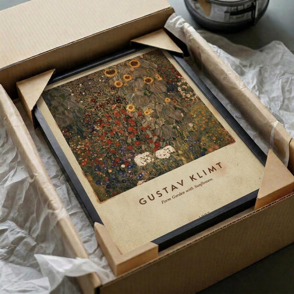 May include: Framed Gustav Klimt reproduction print showing a vibrant garden scene with sunflowers and wildflowers in rich greens, yellows, and reds. The artwork sits in a black frame within an open cardboard shipping box surrounded by white tissue paper on a neutral surface. A ceramic bowl is partially visible in the background.