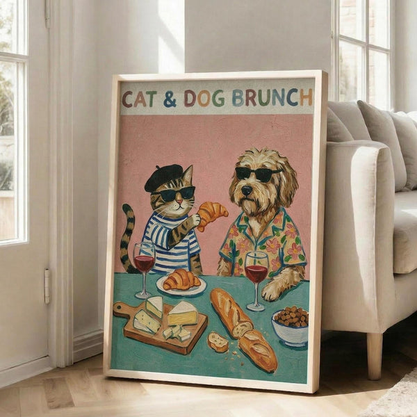 May include: Framed poster print leaning against white wall next to beige sofa in bright living room. Artwork shows anthropomorphic tabby cat in striped shirt and beret alongside shaggy dog in floral Hawaiian shirt, both wearing sunglasses at brunch table with croissants, wine, cheese board, and bread. Pink and teal color scheme with colorful title text at top.