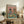 May include: Framed poster showing a tabby cat in beret and striped shirt sharing brunch with a shaggy dog in floral shirt, both wearing sunglasses at turquoise table with croissants, cheese, bread, wine glasses and olives on pink background, displayed on rustic wooden sideboard with books, plant, and ceramic vases in cozy living room setting