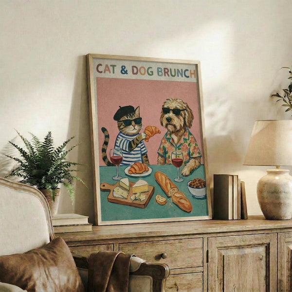 May include: Framed poster showing a tabby cat in beret and striped shirt sharing brunch with a shaggy dog in floral shirt, both wearing sunglasses at turquoise table with croissants, cheese, bread, wine glasses and olives on pink background, displayed on rustic wooden sideboard with books, plant, and ceramic vases in cozy living room setting