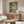 May include: Three vintage Portuguese sardine can labels displayed in a framed green artwork above a mid-century walnut credenza. The labels show "Tricana," "Prata Do Mar," and "Minor" brands with colorful retro designs. Scene includes a white curved chair, brass floor lamp, modern mushroom table lamp, oval glass coffee table, and cream textured rug in a minimalist living room with white walls.