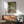 May include: Three vintage Portuguese sardine can labels displayed in a wooden frame with green matting, hanging above a mid-century modern wooden sideboard. The labels show Tricana, Prata Do Mar, and Minor brands with colorful retro graphics. A sage green upholstered armchair with wooden arms sits nearby, with a white ceramic table lamp and round wooden coffee table completing the scene. Warm natural lighting creates shadows on white walls.