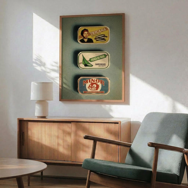 May include: Three vintage Portuguese sardine can labels displayed in a wooden frame with green matting, hanging above a mid-century modern wooden sideboard. The labels show Tricana, Prata Do Mar, and Minor brands with colorful retro graphics. A sage green upholstered armchair with wooden arms sits nearby, with a white ceramic table lamp and round wooden coffee table completing the scene. Warm natural lighting creates shadows on white walls.