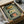 May include: Three vintage-style Portuguese food labels displayed in a green matted frame inside an open cardboard box with tissue paper. The rectangular labels feature Tricana sardines with a woman's portrait, Compadrio sardines with green leaf design, and Minor brand with red logo. Frame appears dark colored against olive green matting, photographed as an unboxing scene with wooden handles visible and white tissue paper packaging.